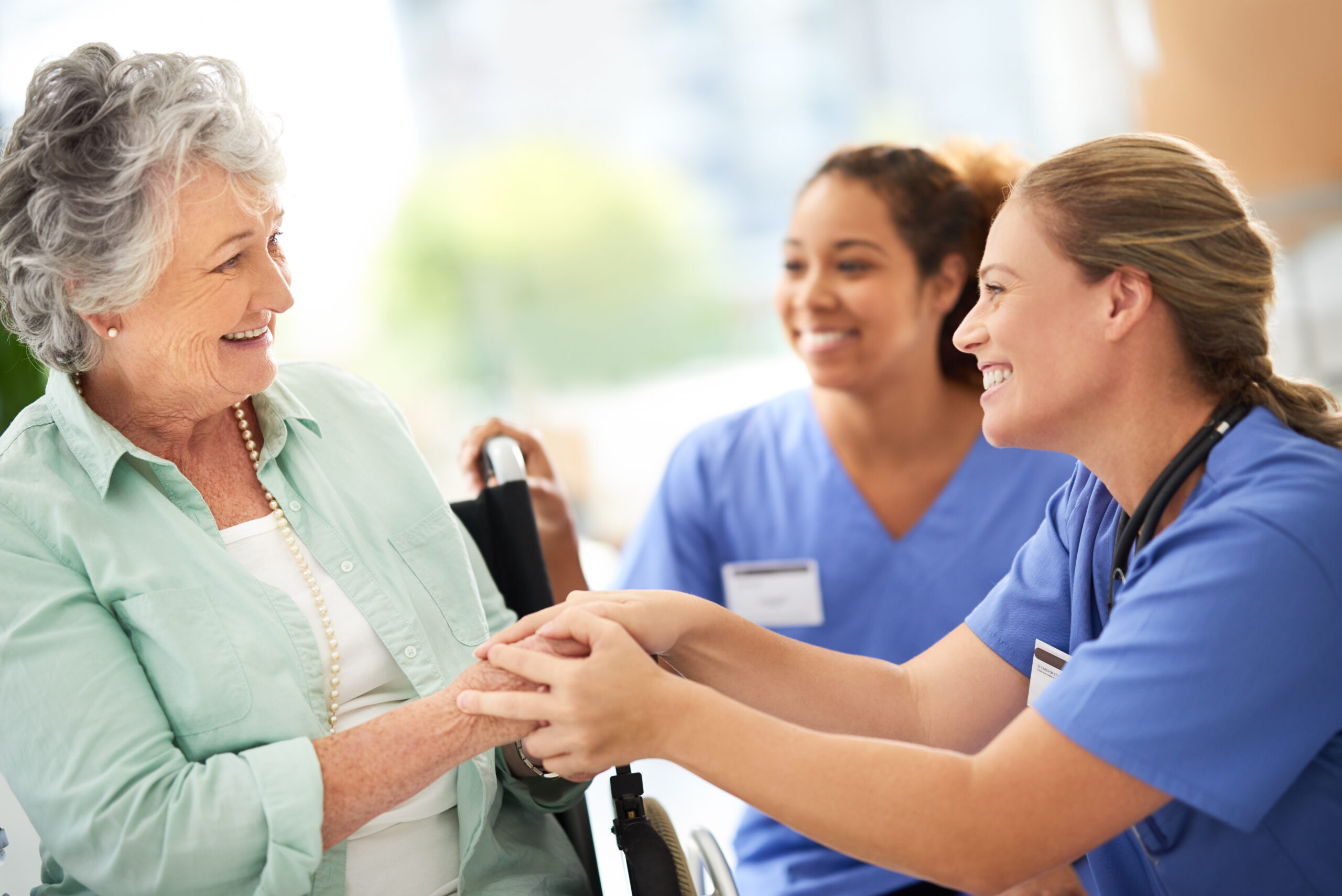 A hospice nurse holding the hand of an older woman in a wheelchair while a second caregiver smiles beside them